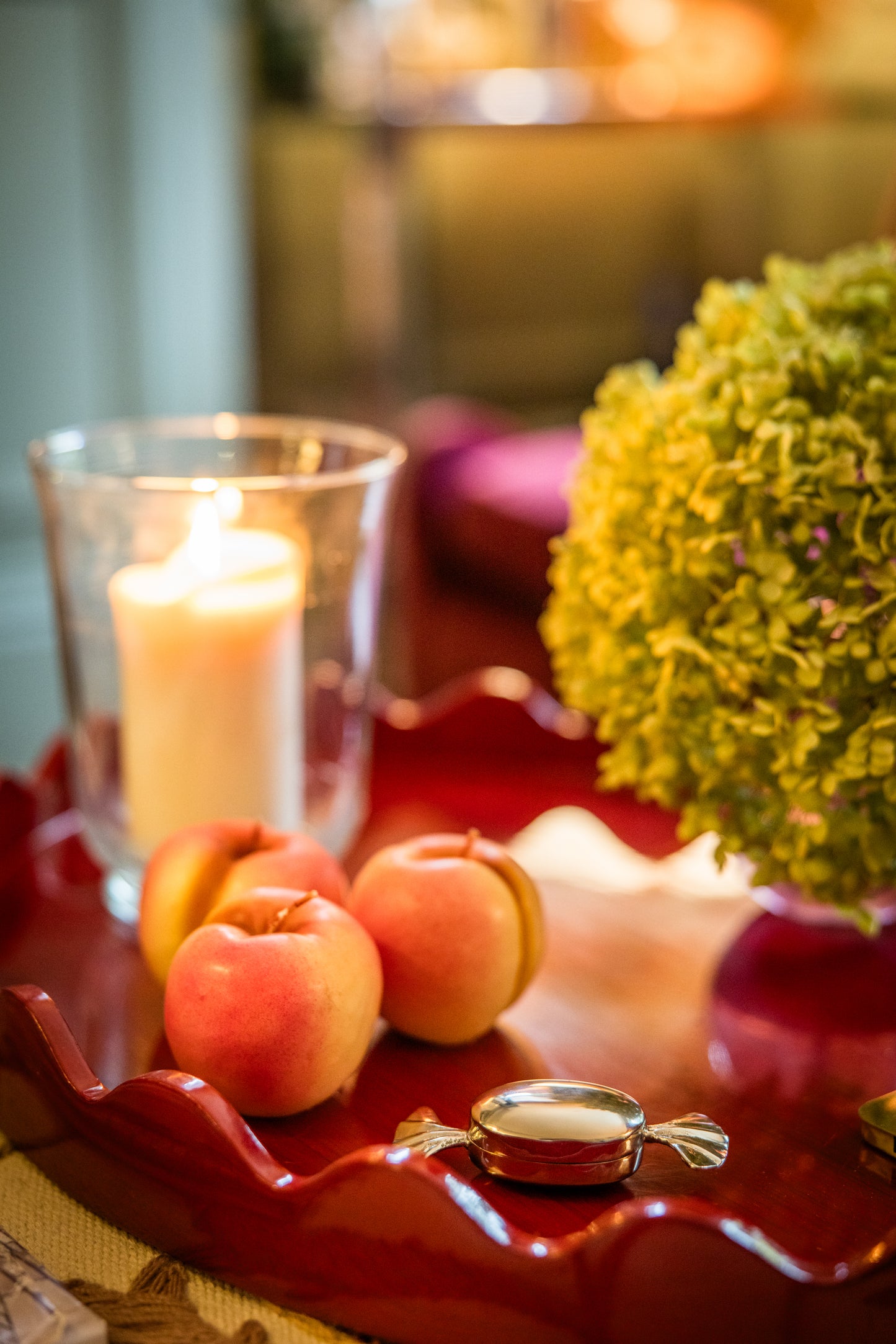 Decorative side table with ric rac edge trim. On top is a hard candy shaped box, decorative apples, a candle in a glass shade, and hydrangea flowers on a red tray.