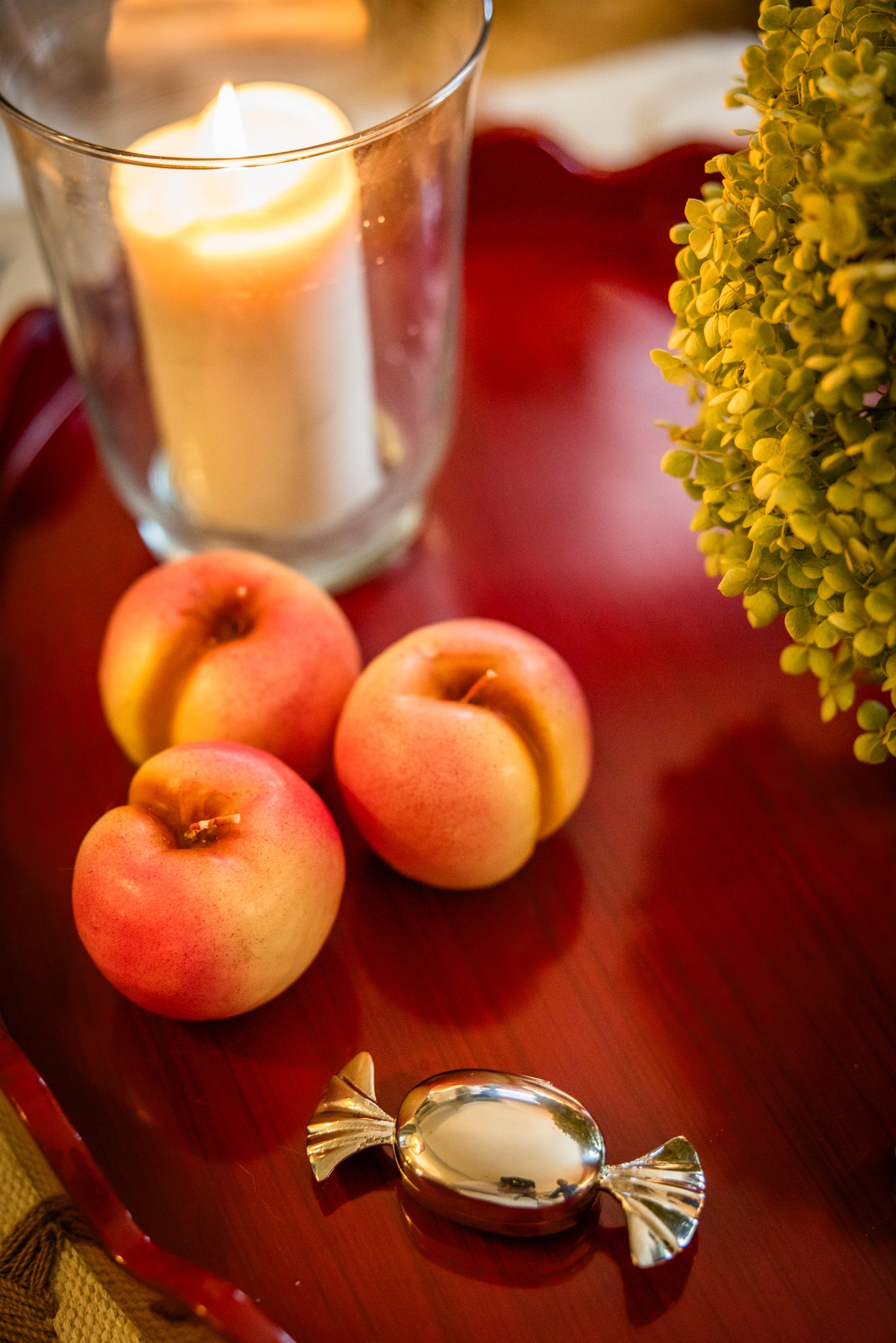 Nectarine shaped candles, a large column candle in a hurricane glass shade, and a small chrome candy shaped decorative container on a red wood tray.