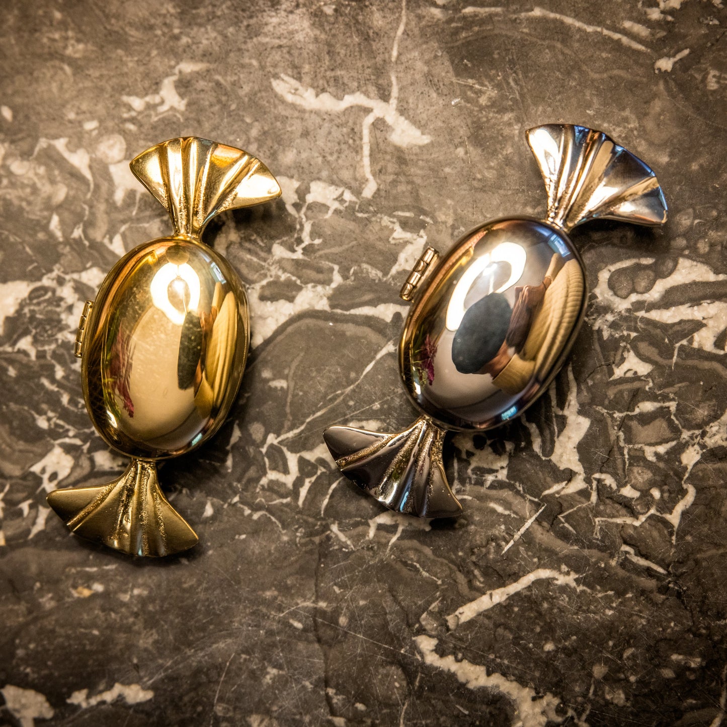 A gold and a chrome candy-shaped hinged storage container on a dark marble surface.