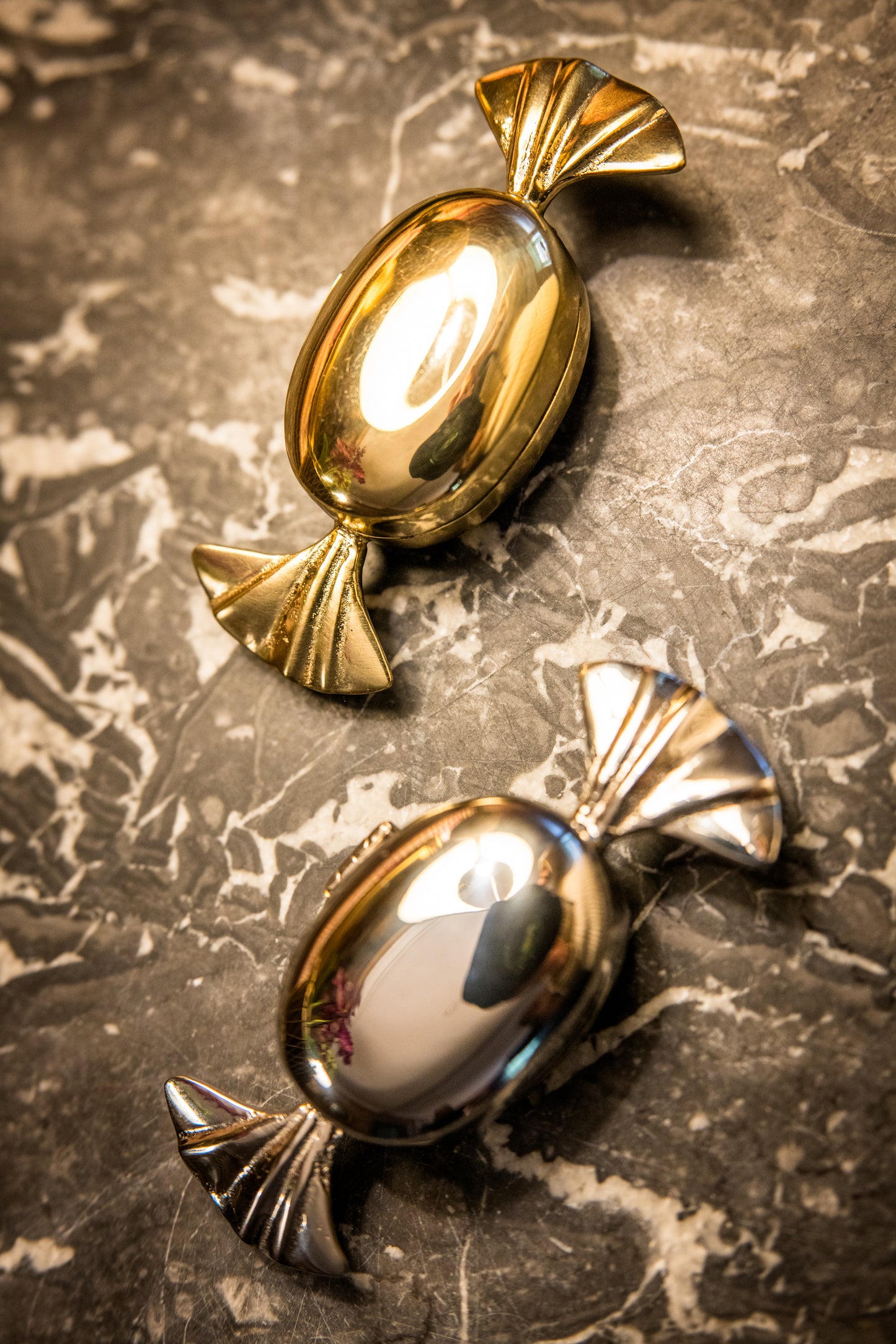 Two metallic candy-shaped decorative objects one gold the other silver on a textured dark grey and white marble surface.