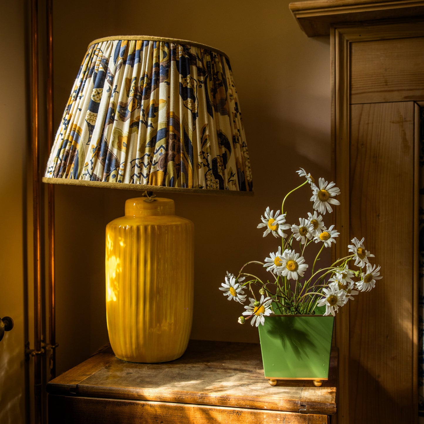 Yellow lamp with patterned shade on a wooden surface next to a green metal vase with white daisy flower arrangement.