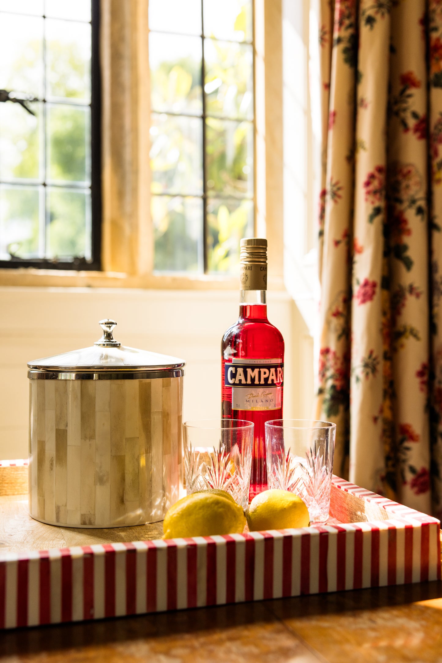 Bottle of Campari with ice bucket, crystal tumbler glasses and lemons on a tray by a window with floral curtains.