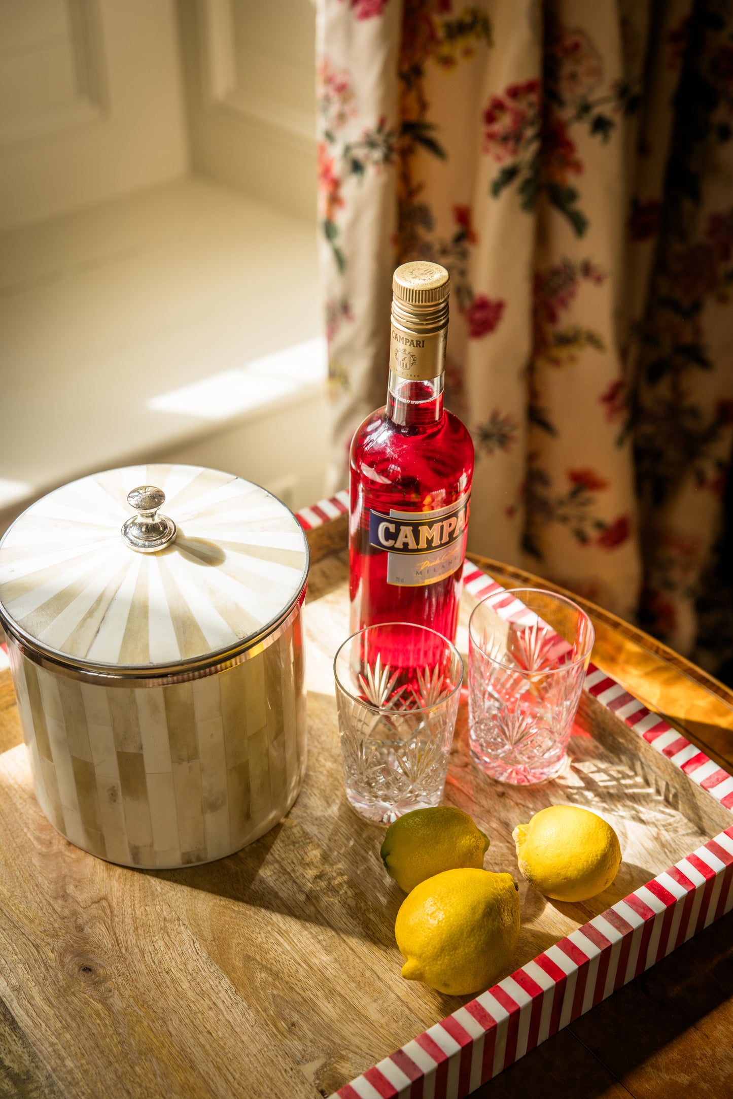 Large drinks tray with round bone inlay ice bucket, Campari bottle, glasses, and lemons on a wooden surface with floral curtains in the background.