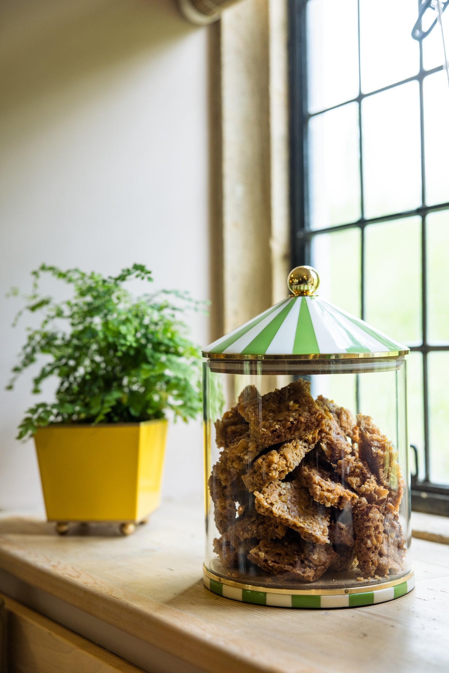 Glass jar with green stripe lid containing flapjacks on a windowsill with a square houseplant in the background