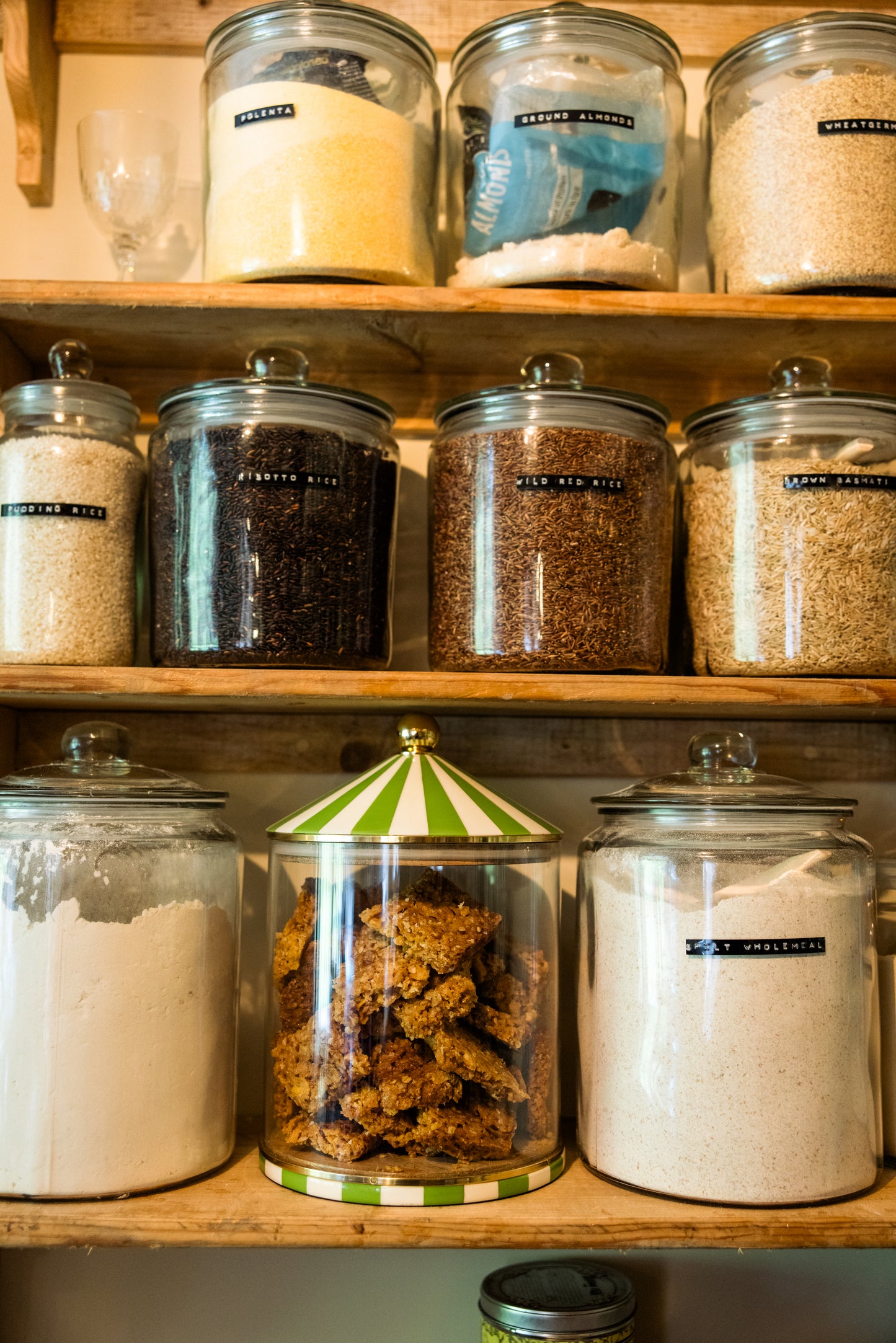 Pantry larder shelves with large glass jars containing various food items including flour, rice, oats on a wooden shelf. All but one container is traditional, the middle one on the lowest shelf is a green and white circus design.