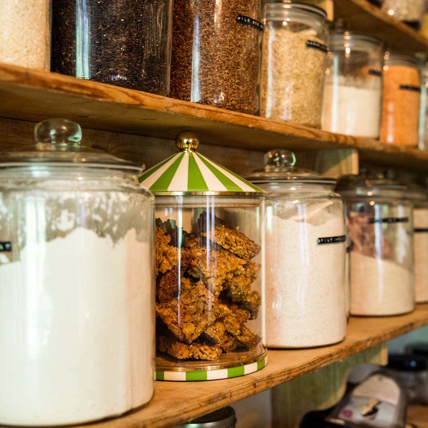 Shelves with various large food storage jars containing different food items in a kitchen cupboard, one stands out with a unique green circus top design.