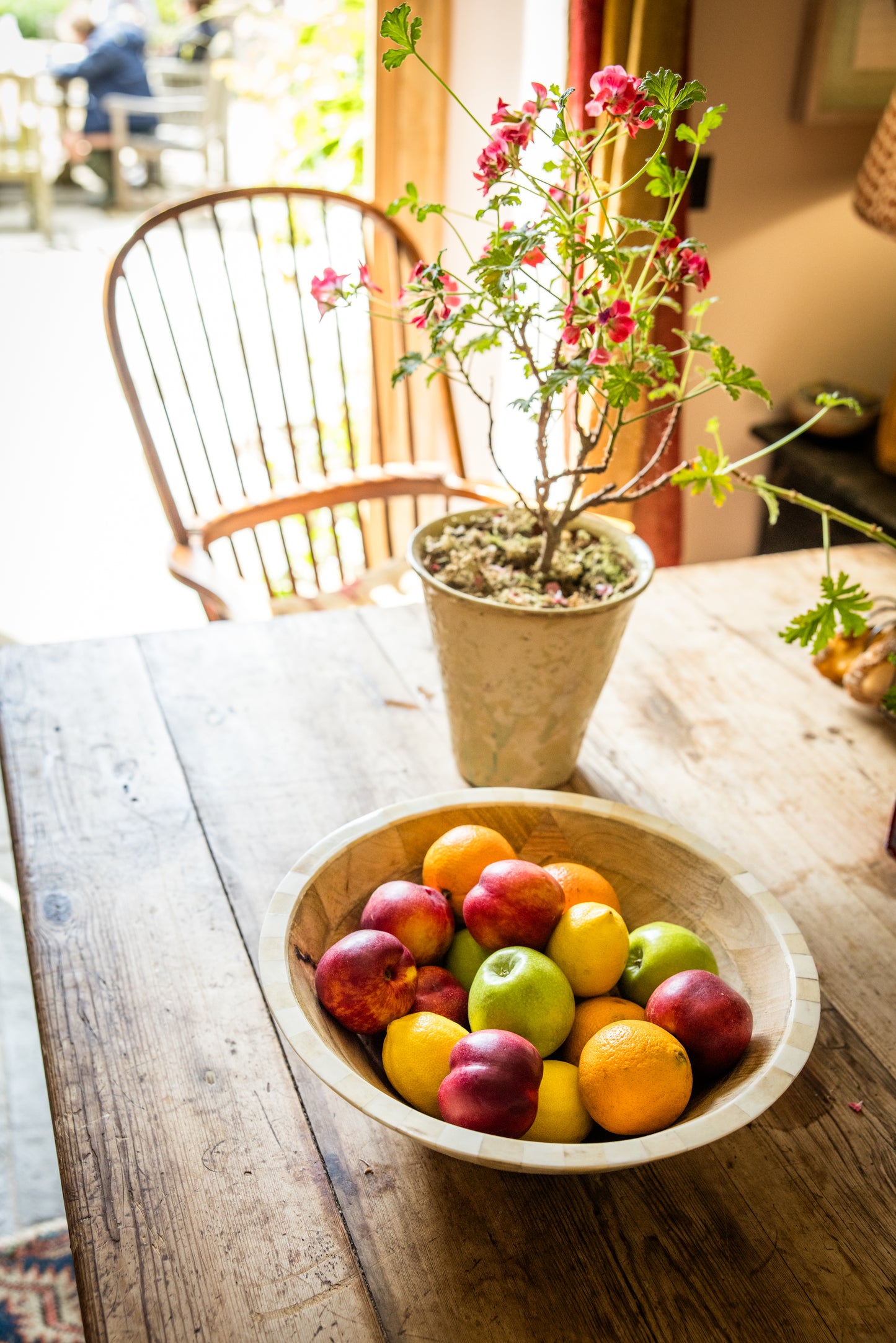 Bowl of fruits on a dining table with a potted plant and wooden armchair in the background.