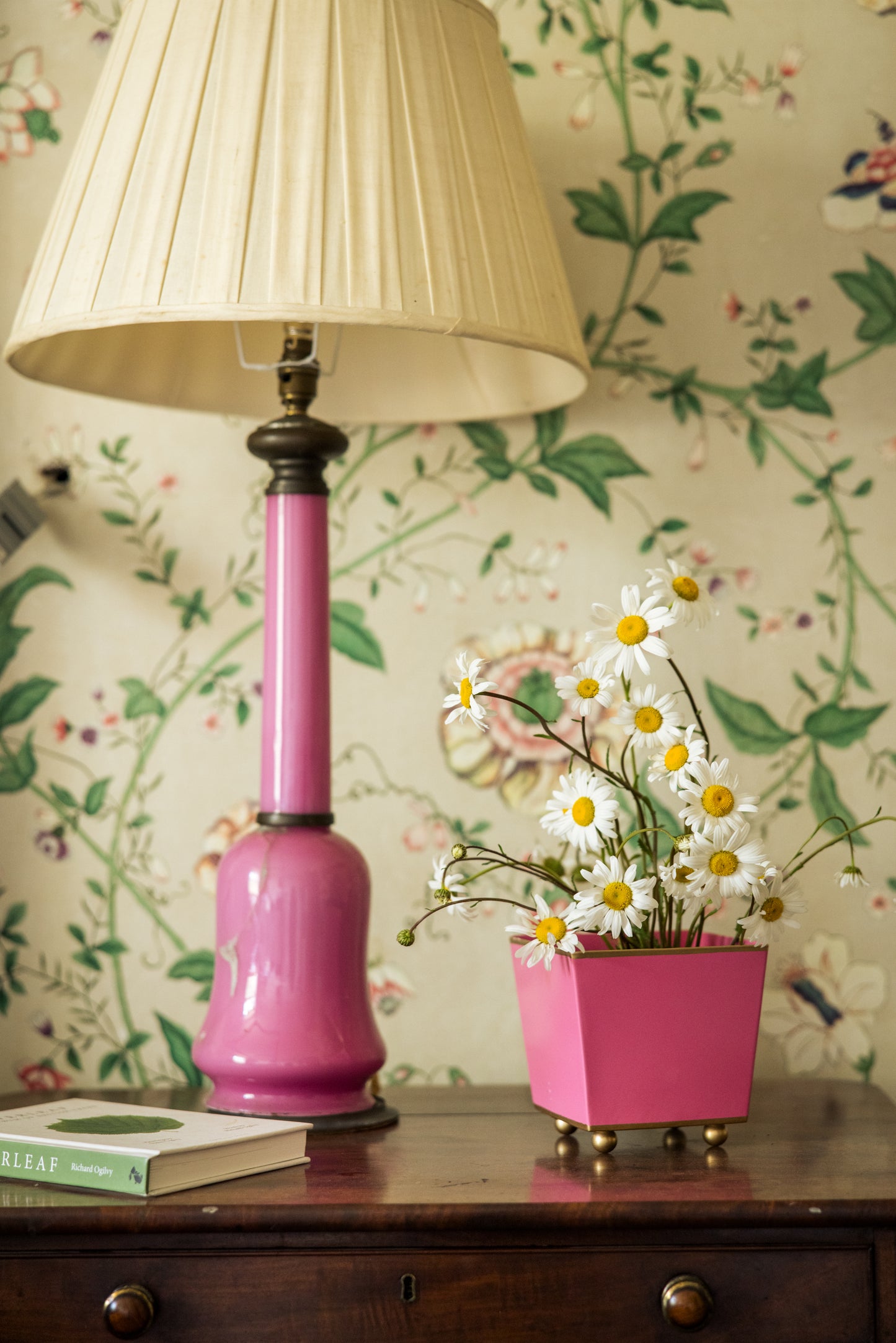 Matching pink lamp and plant pot with daisies on a wooden table against floral wallpaper.