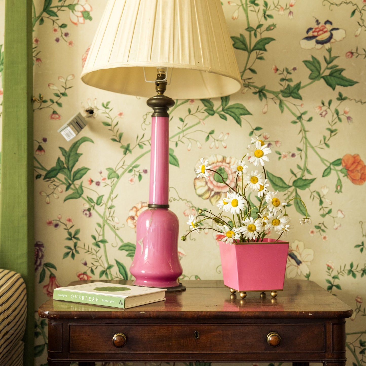 Pink lamp and square shaped flowerpot with daisies on a wooden side table against botanical floral wallpaper