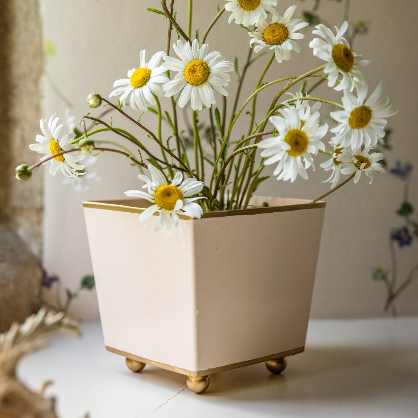 Off white toleware planter with gold edge and ball feet filled with daisies on a neutral background.
