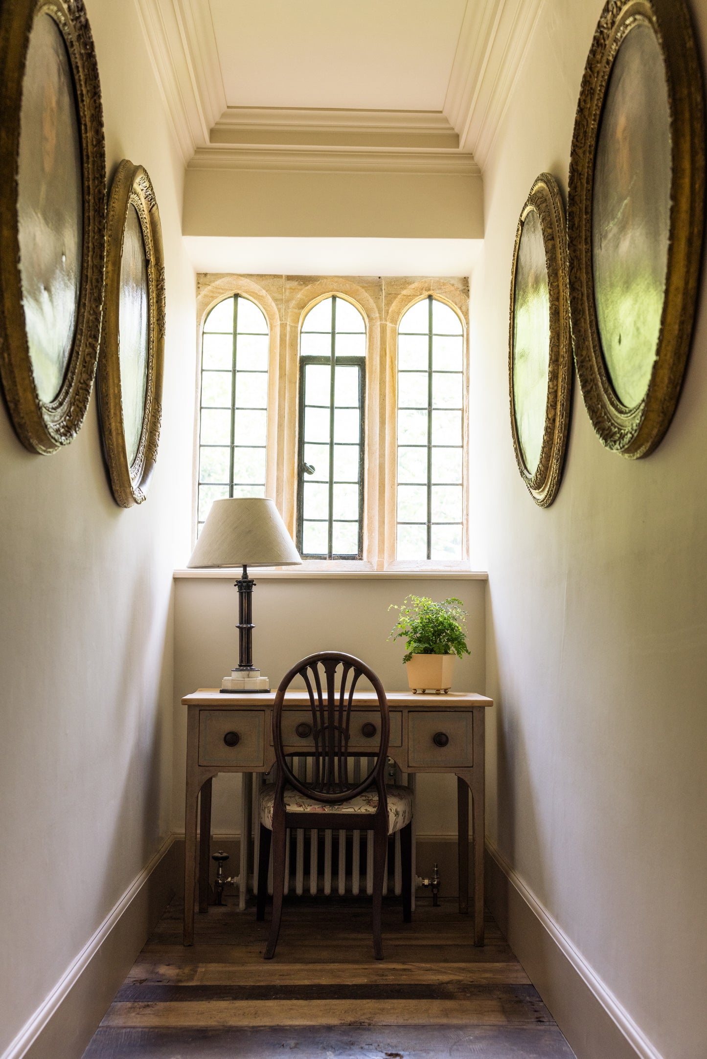 Narrow hallway with a desk, chair, lamp and houseplant at the end, the walls decorated with round paintings.