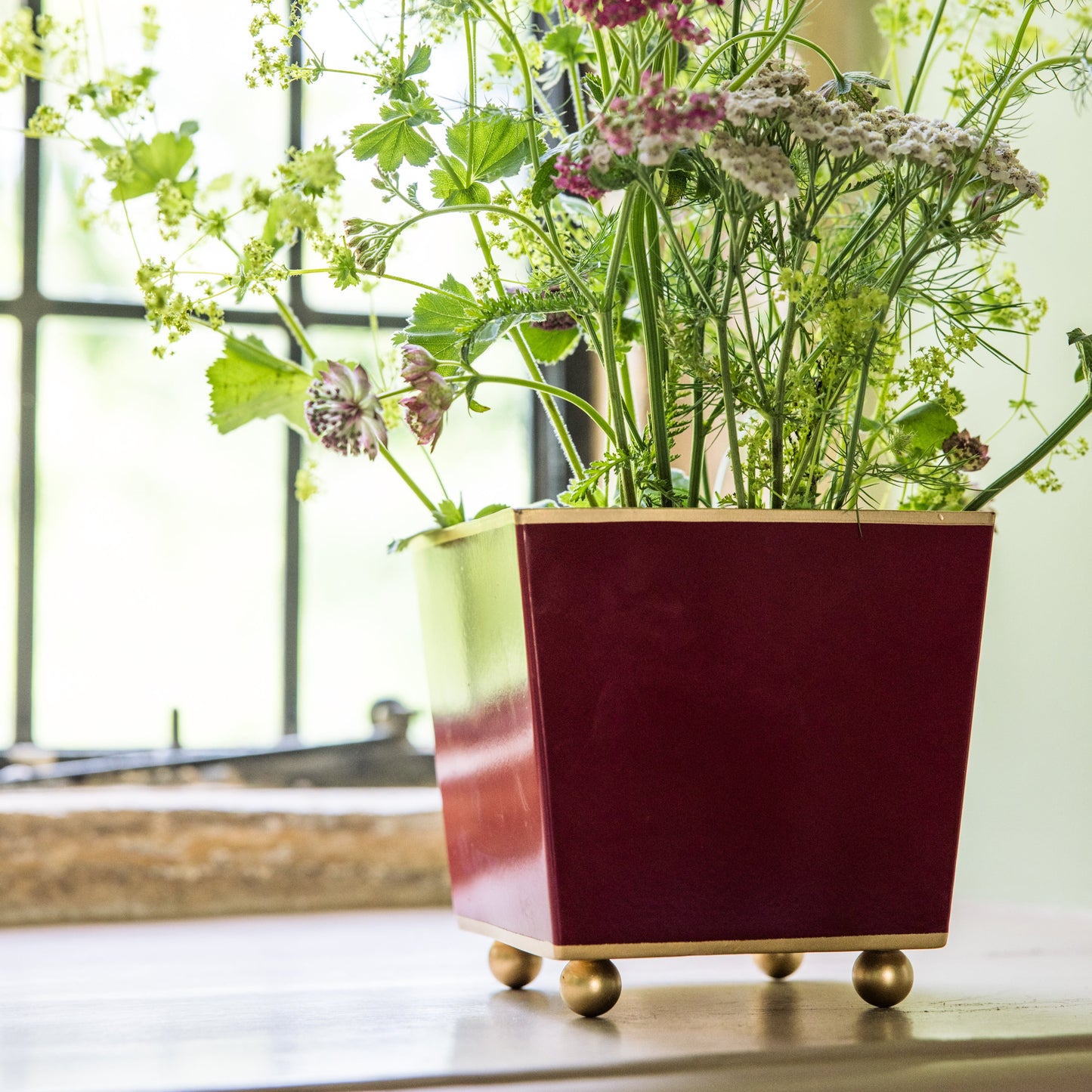 Decorative burgundy planter with gold ball feet and flowers inside on a windowsill.