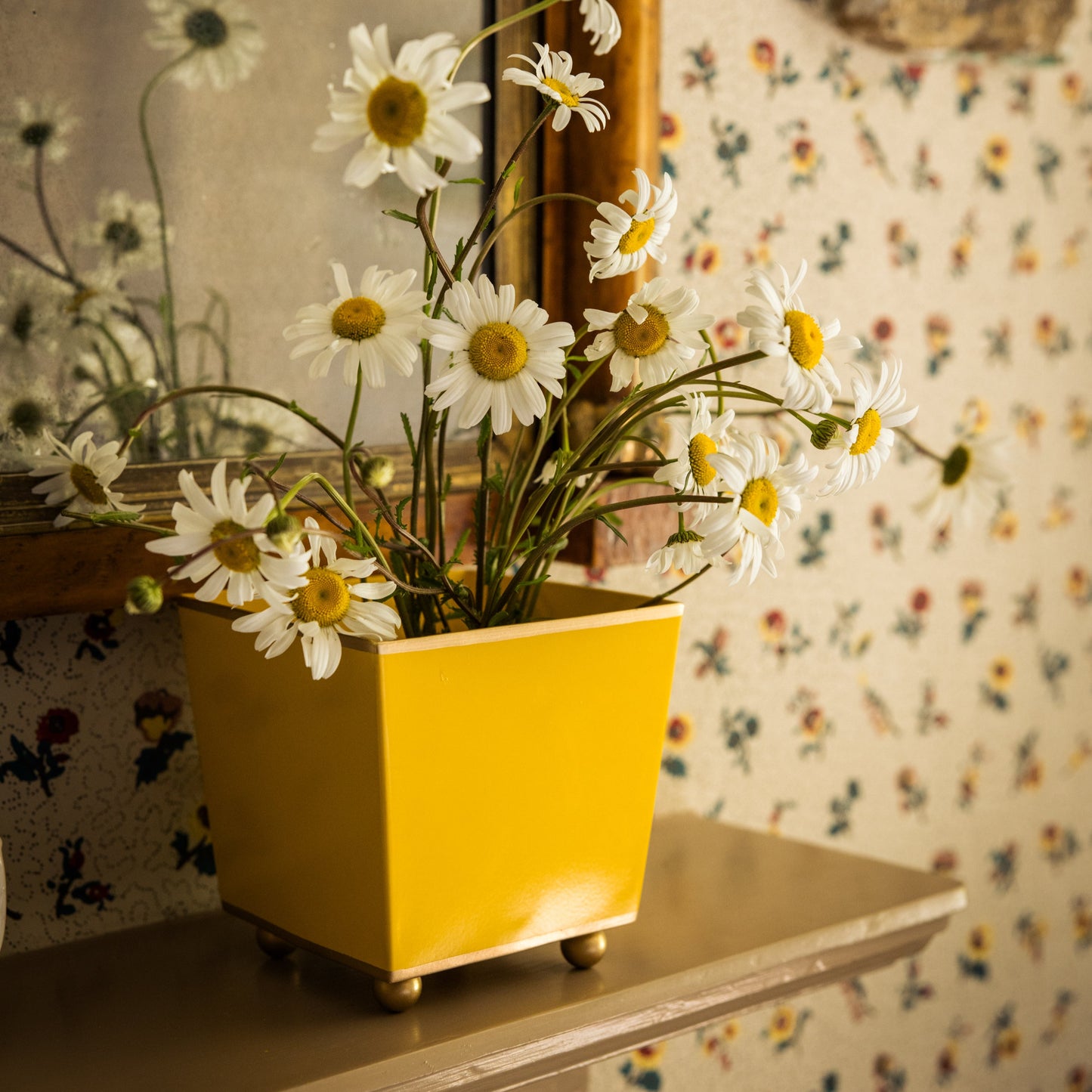 Yellow flowerpot with daisies on a mantlepiece against a floral wallpapered wall and mirror in the background.