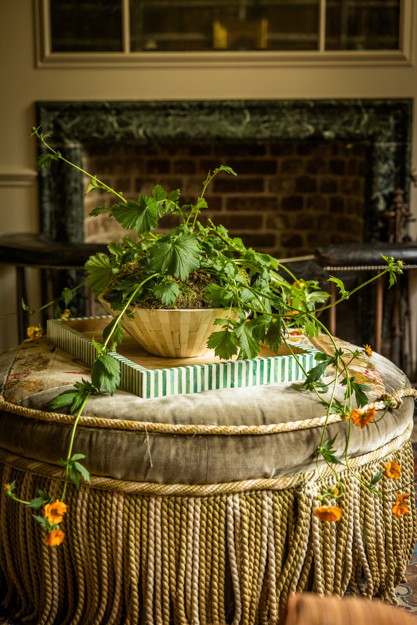 Potted plant on a large decorative tray resting on a rope tassel round ottoman with a fireplace in the background