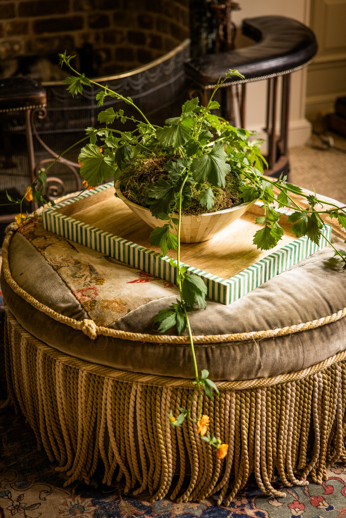 Round tassel ottoman with a plant on top of a tray in a cozy room setting with a fireplace in the background