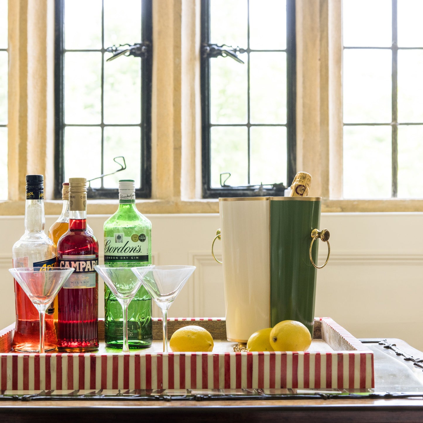 Alcohol bottles, martini glasses and a wine cooler on a striped tray with lemons in front of a window.