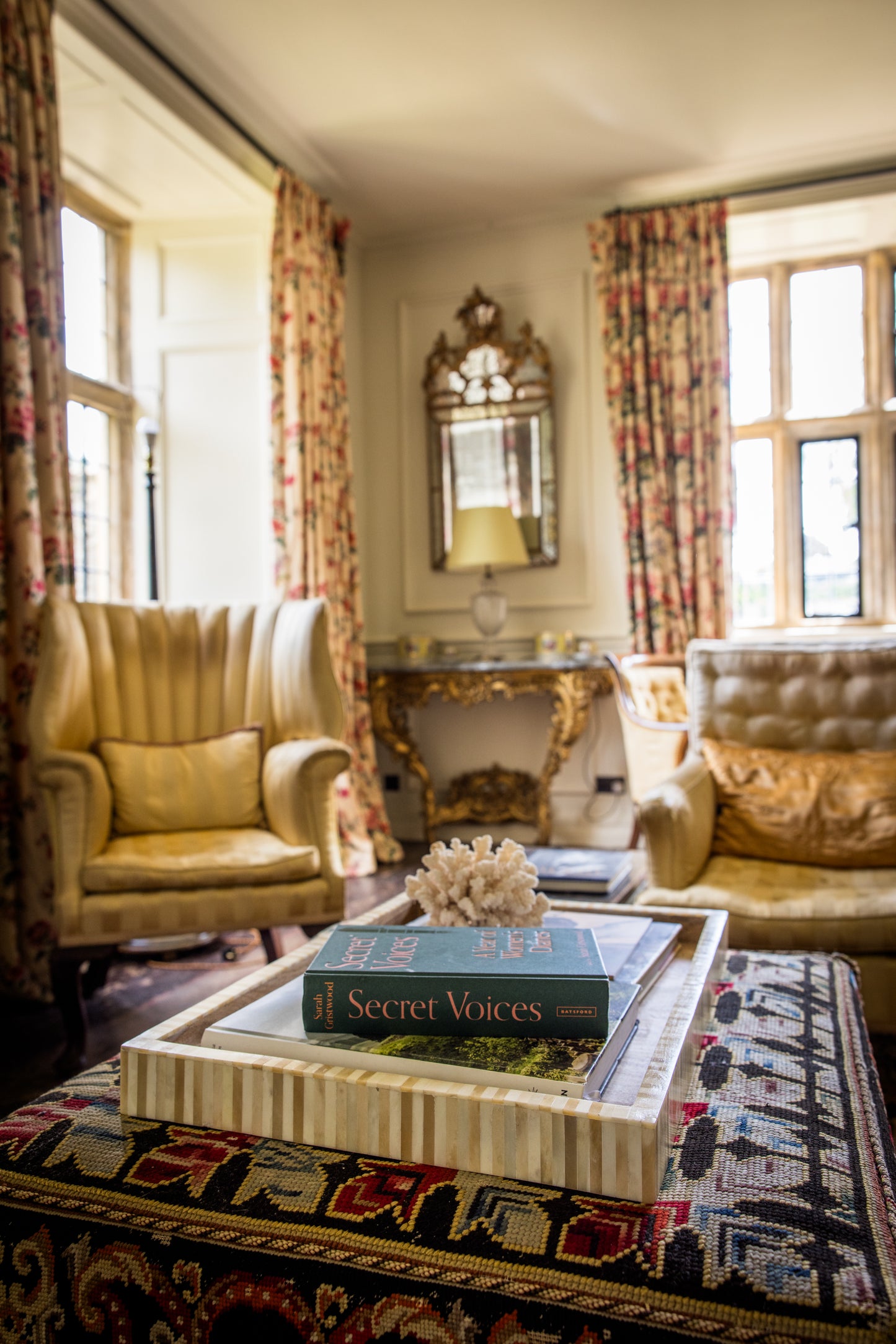 Beige striped tray organising a coffee table book display on an ottoman in a luxurious living room with ornate furniture in the background.