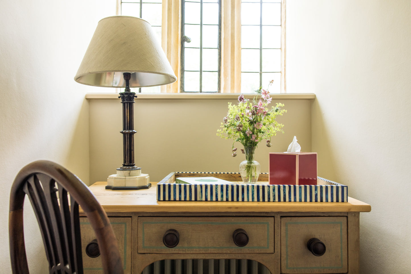 Wooden desk with lamp next to flowers, a book and a tissue box in a room with a window.