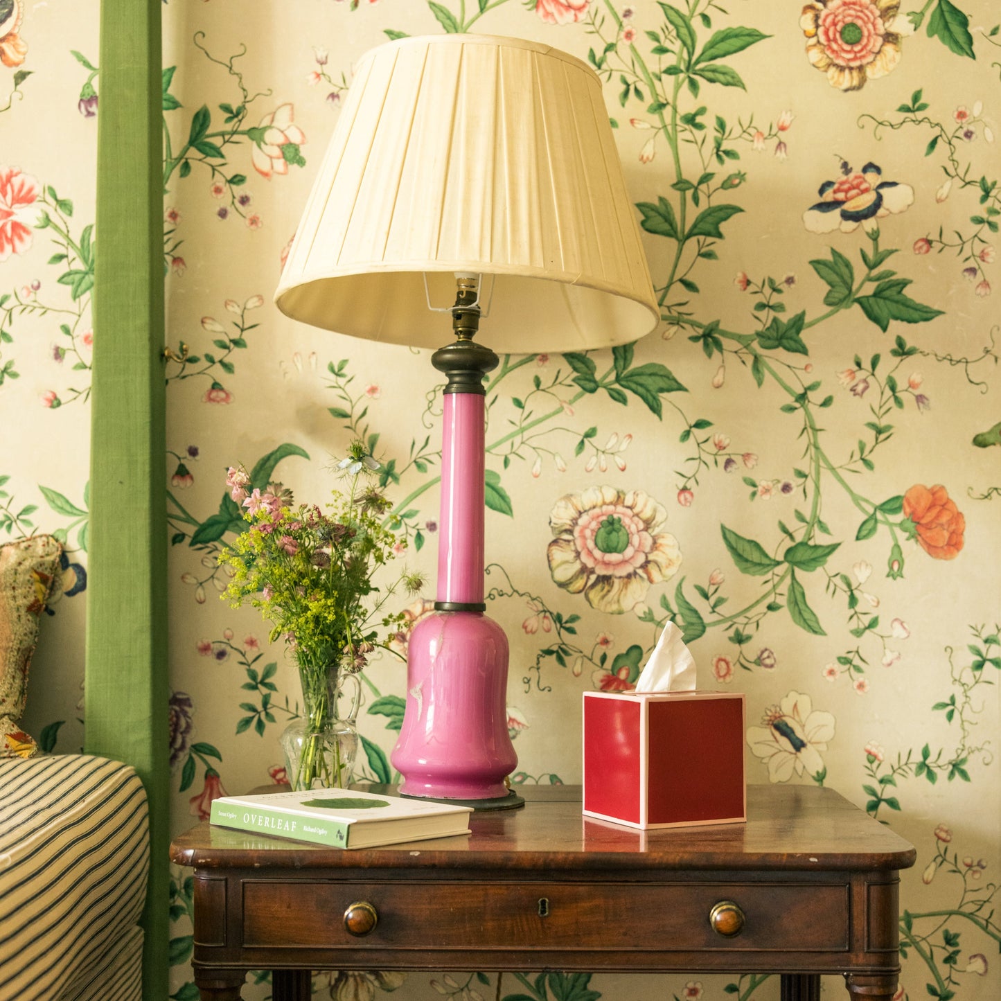 Red tissue box on a bedside table next to a pink lamp, flowers and book against floral botanical wallpaper.