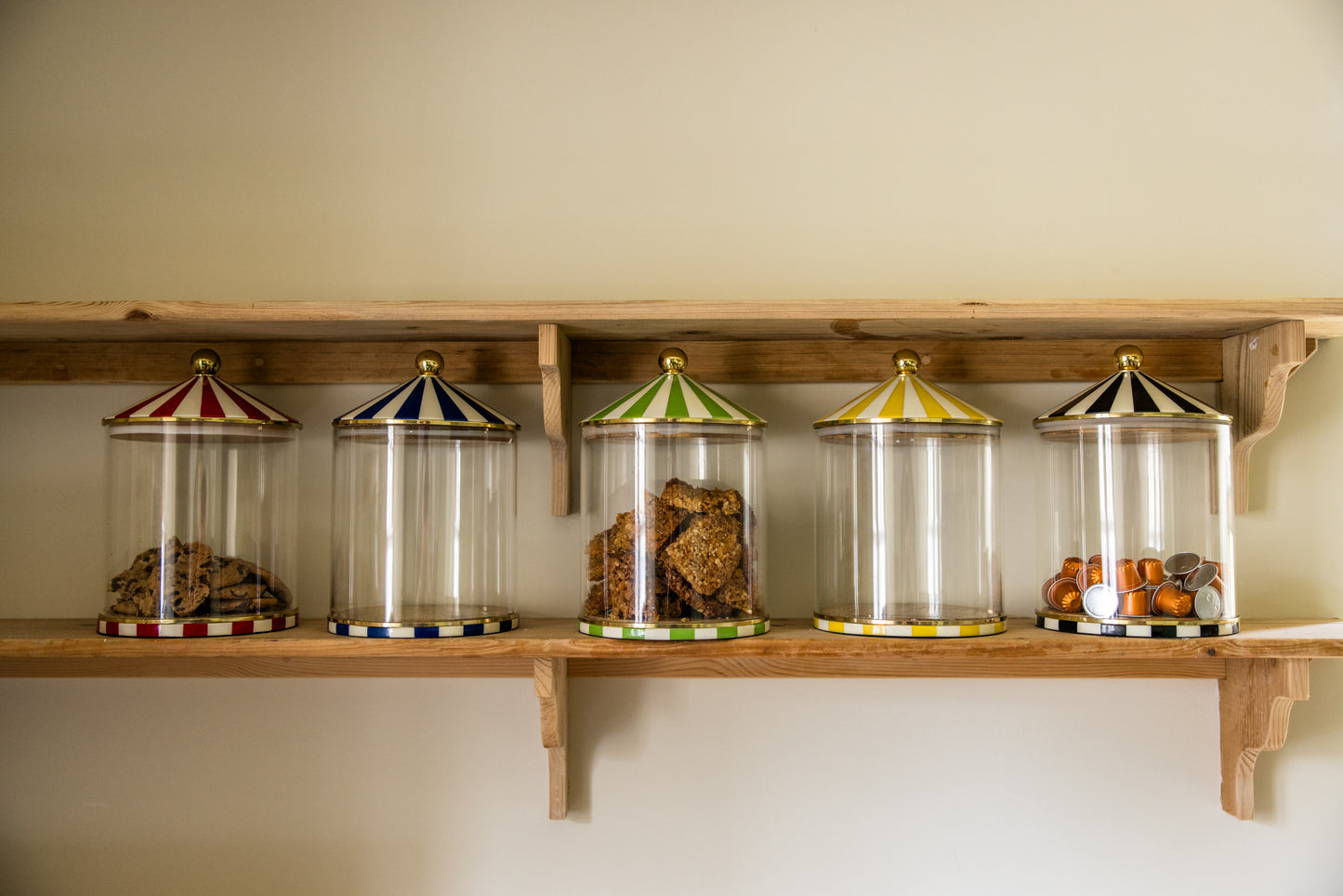 Five transparent cylindrical containers with striped lids on a wooden shelf against a beige wall.