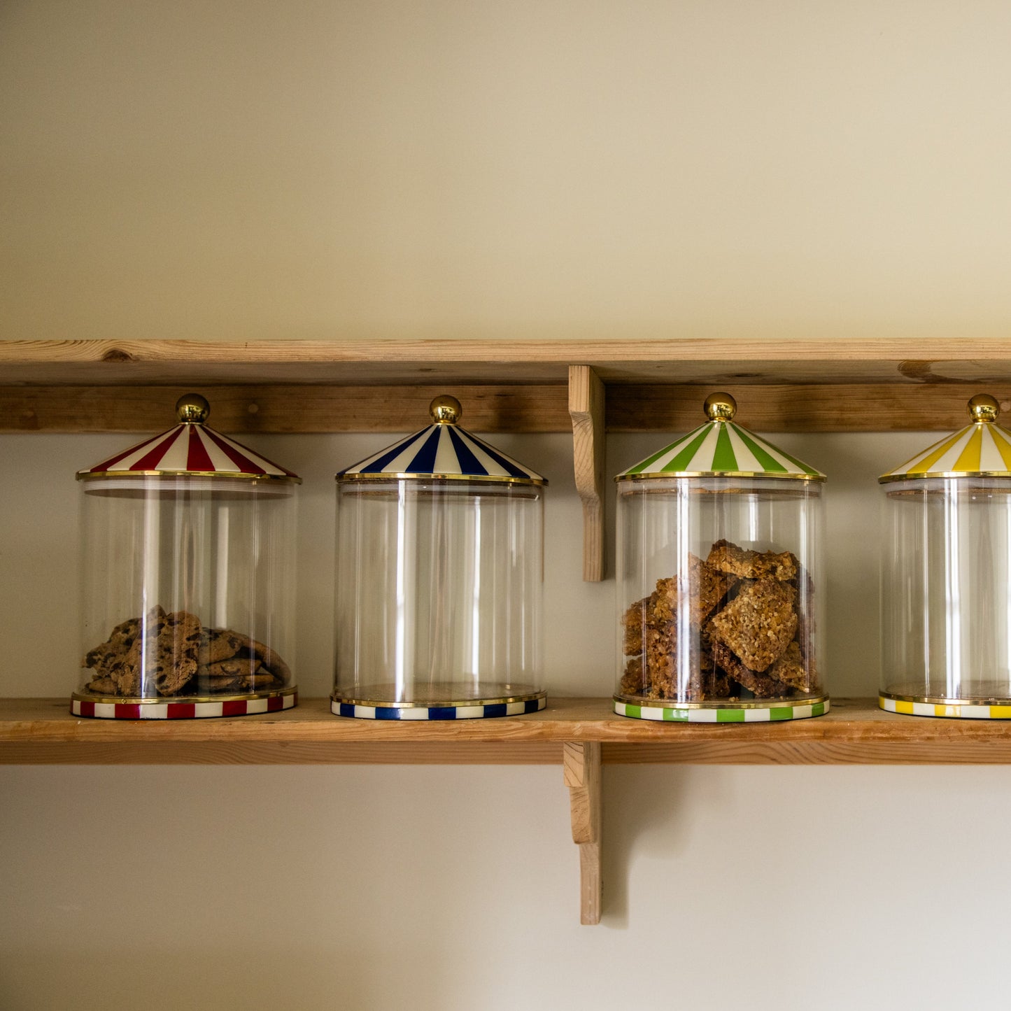 Four glass jars with circus top pitched lids filled with sweet treats on a wooden shelf against a beige wall.