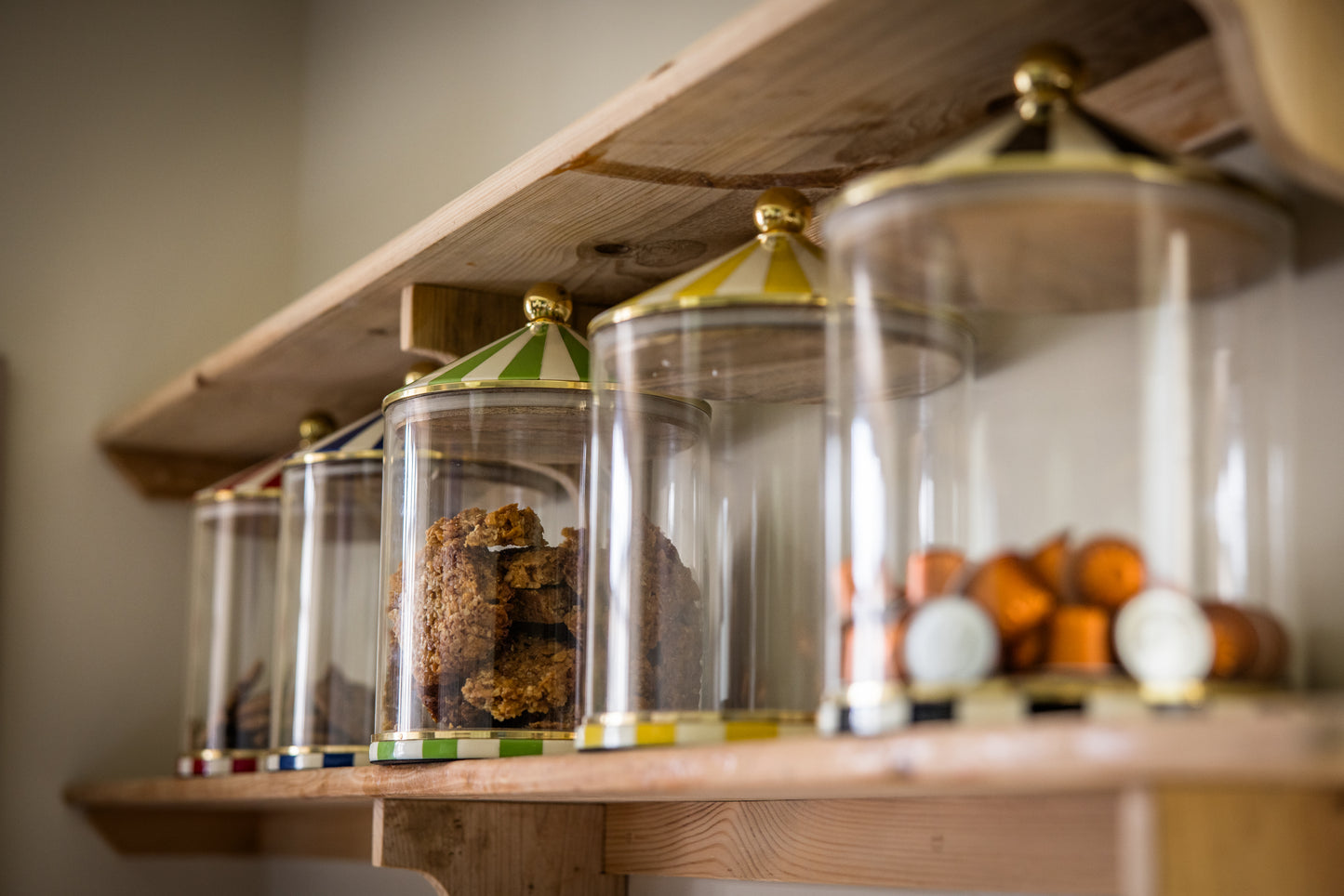 Five colourful glass jars with decorative striped lids on a wooden shelf holding coffee pods, flapjacks and cookies.