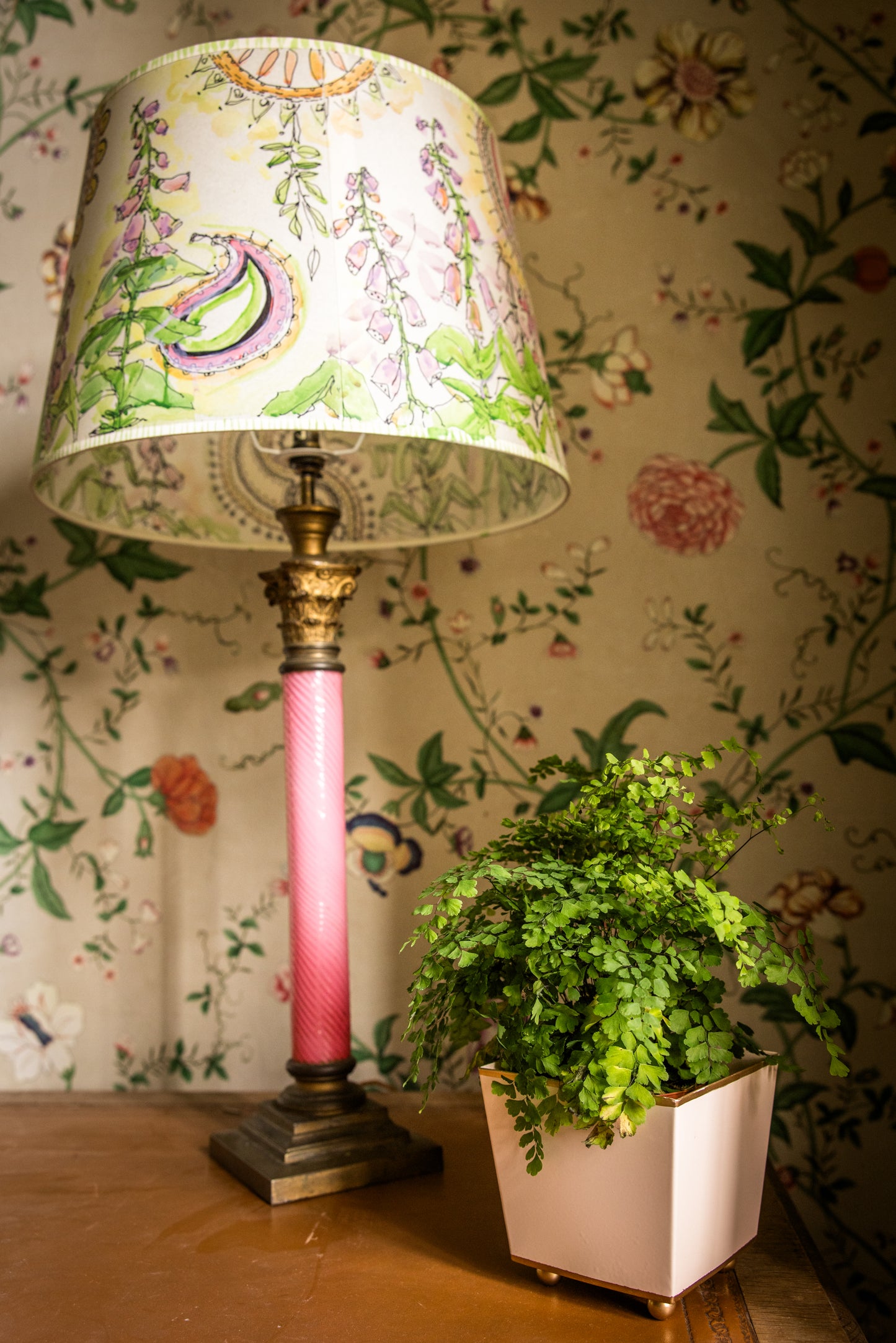Lamp with a floral shade and pink base next to a nude peach colour potted plant pot against a floral wallpapered wall.