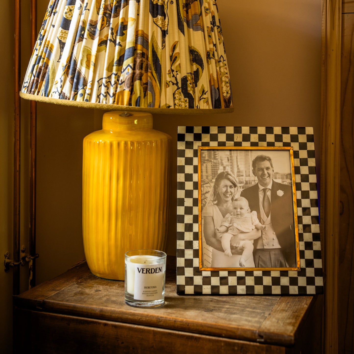 Yellow lamp with patterned shade on a wooden sidetable next to a black and white framed photo and a votive candle.
