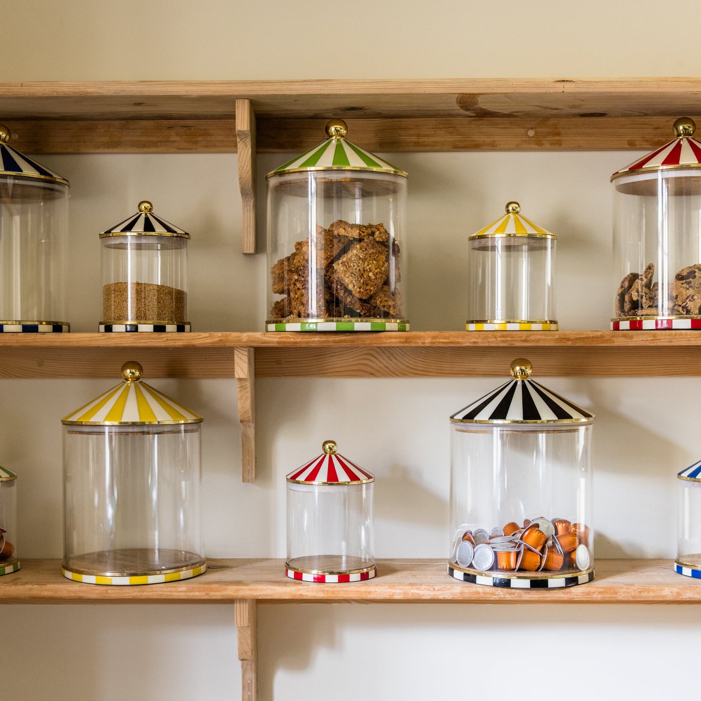 Wooden shelves with glass storage jars in two sizes featuring colourful circus lids on a neutral background.