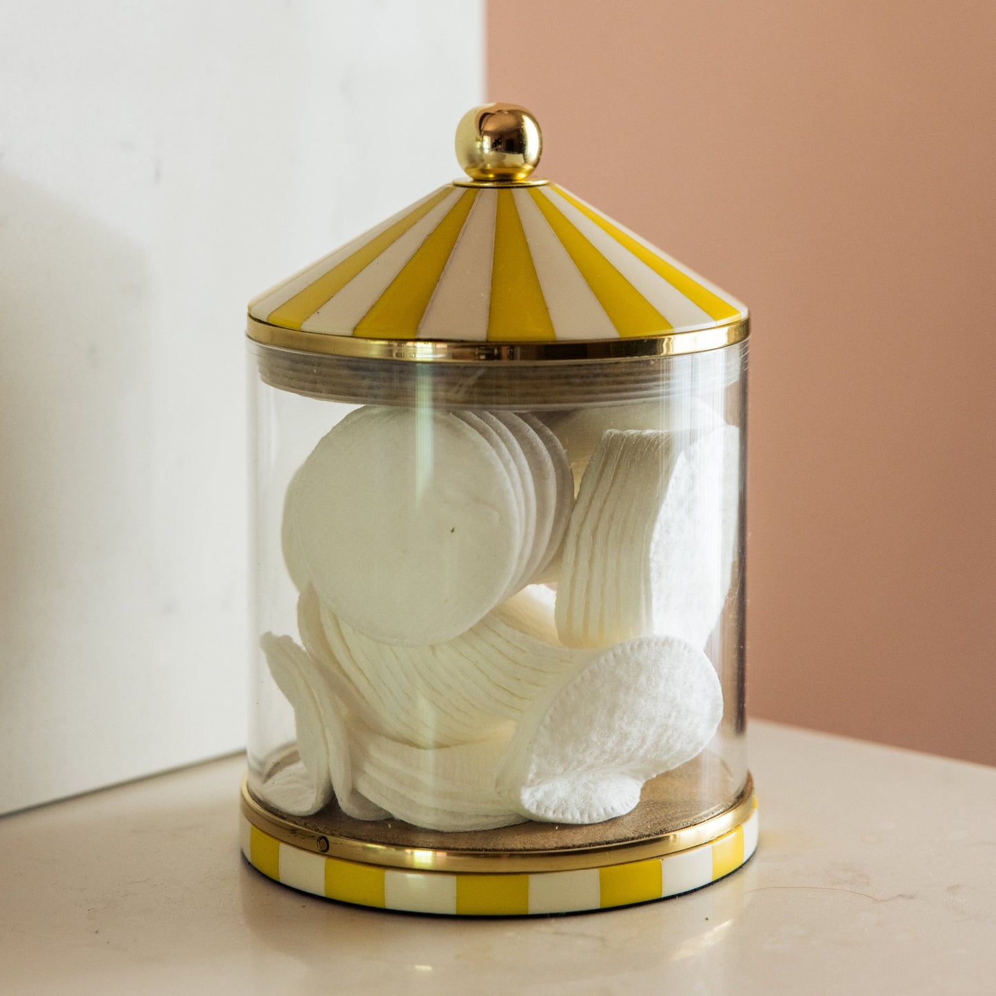 Decorative jar with yellow circus striped lid storing cotton pads on a bathroom shelf.