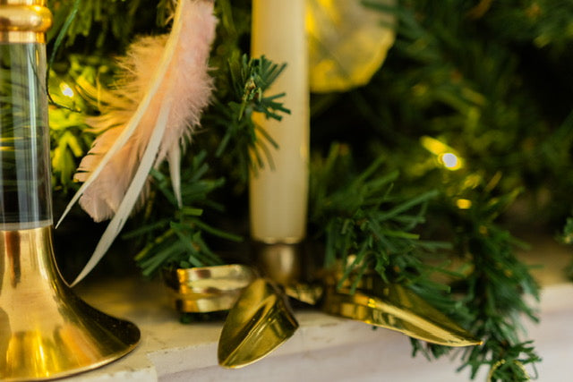 A close up of two candleholders, to the left a traditional brass candlestick with translucent stem, to the right a brass bow with white candle. The background is fairy lights and green foliage.