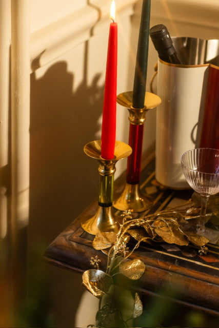 Two classic candlesticks on a table, one is short with a green stem and red lit candle, the other taller with a red stem and black candle next to a wine cooler chilling white wine, gold decorations and a glass.