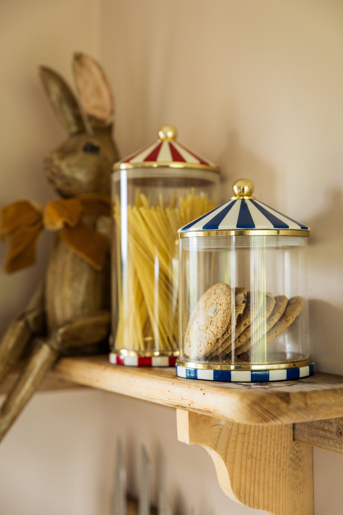 Two circus style glass food storage jars on a shelf with a wooden bunny rabbit in the background. the front one is blue and white stripes, holding cookies and is shorter, behind it is a taller red and white version holding spaghetti.