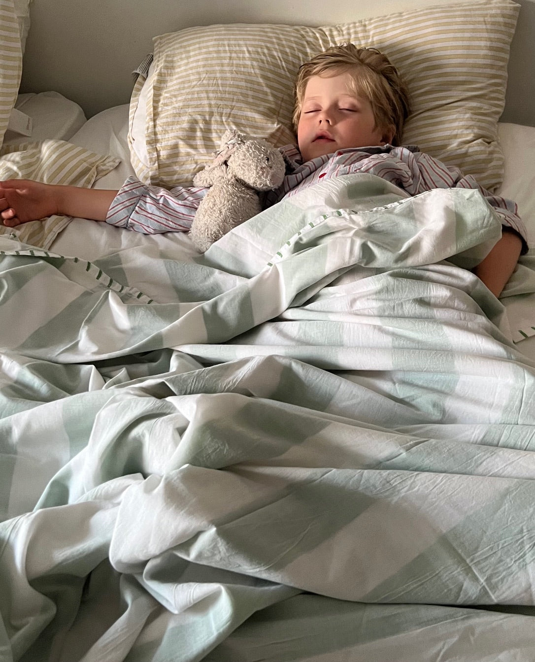 A blonde child sleeps in a bed with an old stuffed animal under a pale green and white cotton bedsheet with contrasting stitching on the edge.