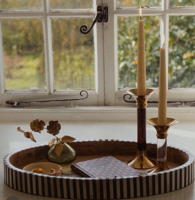 An oval shaped tray on a windowsill with two candlesticks and taper candles lit inside next to a book, vase with metal flower and trinket dish.