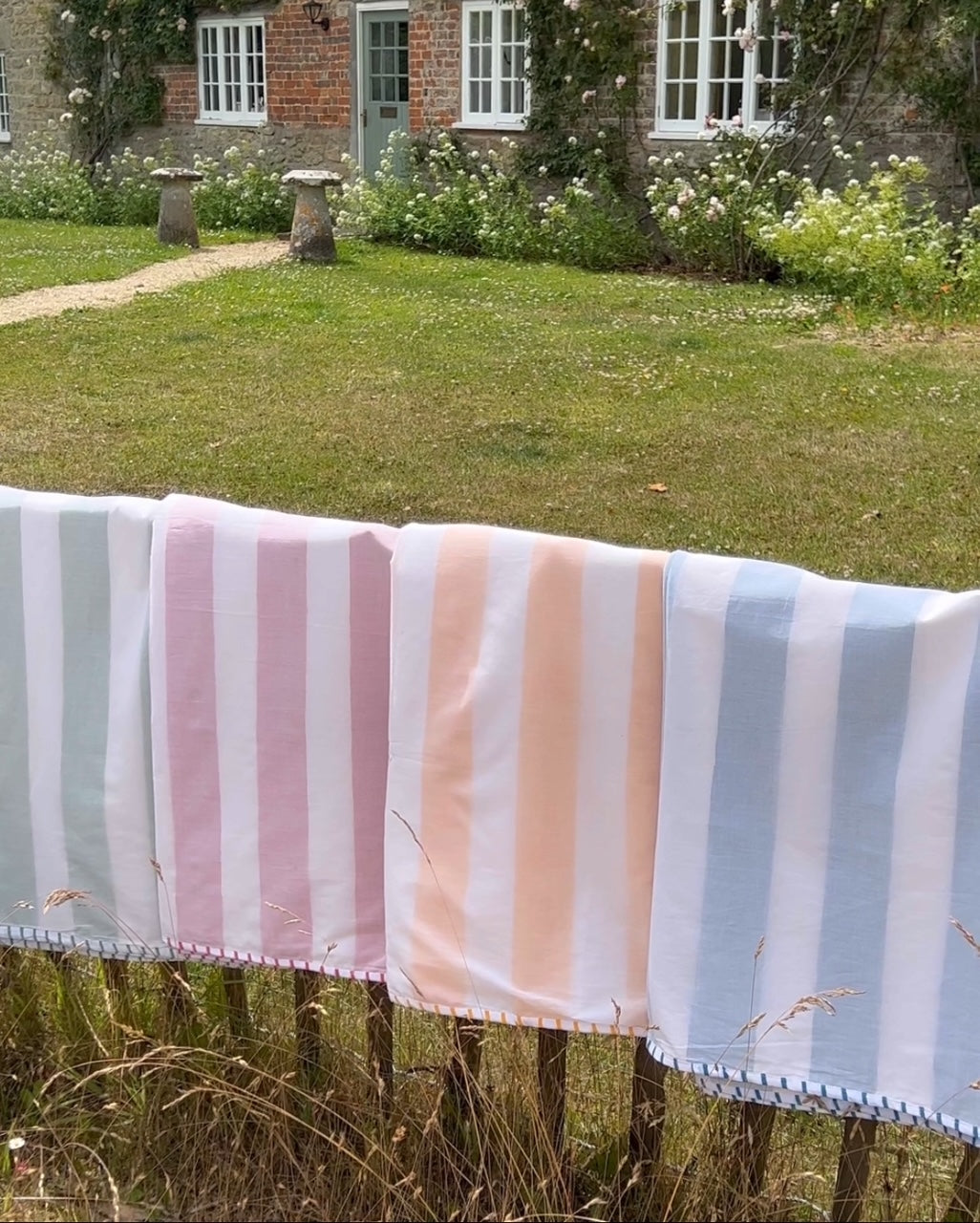 Four striped cotton dohar blankets in pink, blue, green and yellow colours hangs on a wooden fence against an english cottage garden backdrop.