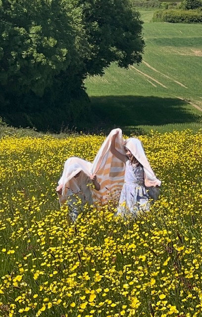 Two children play hiding under an orange and white stripe picnic blanket in a field of yellow buttercups.