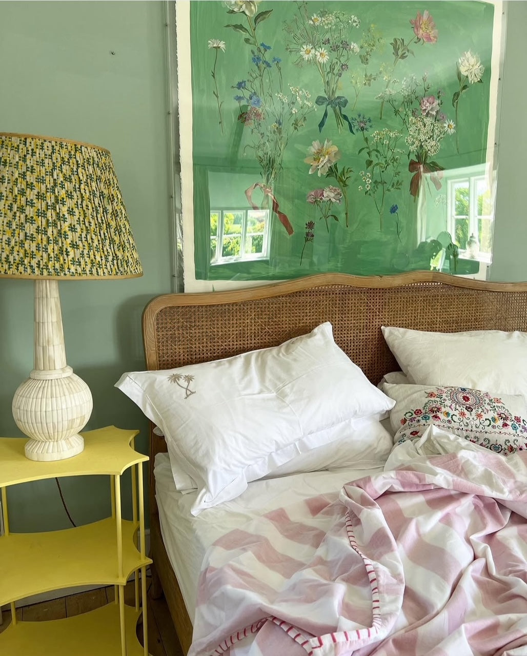 An unmade bed with white pillows, a rattan headboard, embroidered throw pillow and pink stripe bedsheet. The yellow bedside table has a patterned lamp and on the wall behind a green floral artwork reflects bright sunlight through windows.