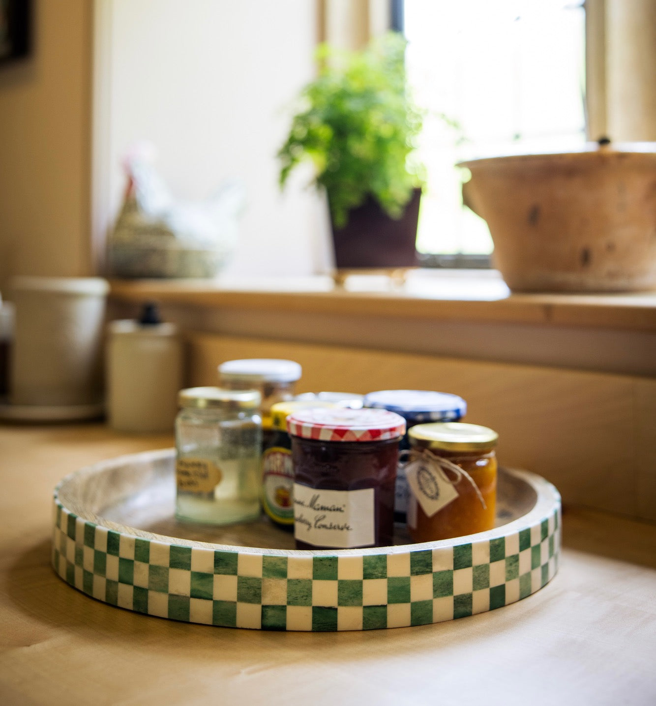 Green checkerboard tray with jars of preserves on kitchen counter with plants and a plant pot on the windowsill in the background.