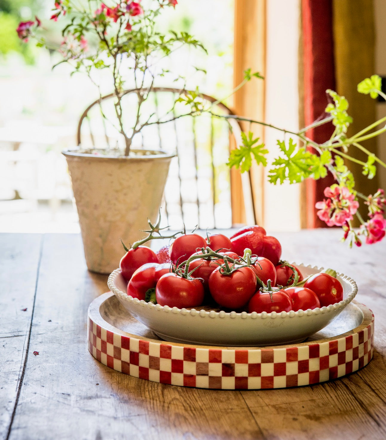 Bowl of tomatoes displayed on a red checkered tray with flowerpot, chair and garden in the background.