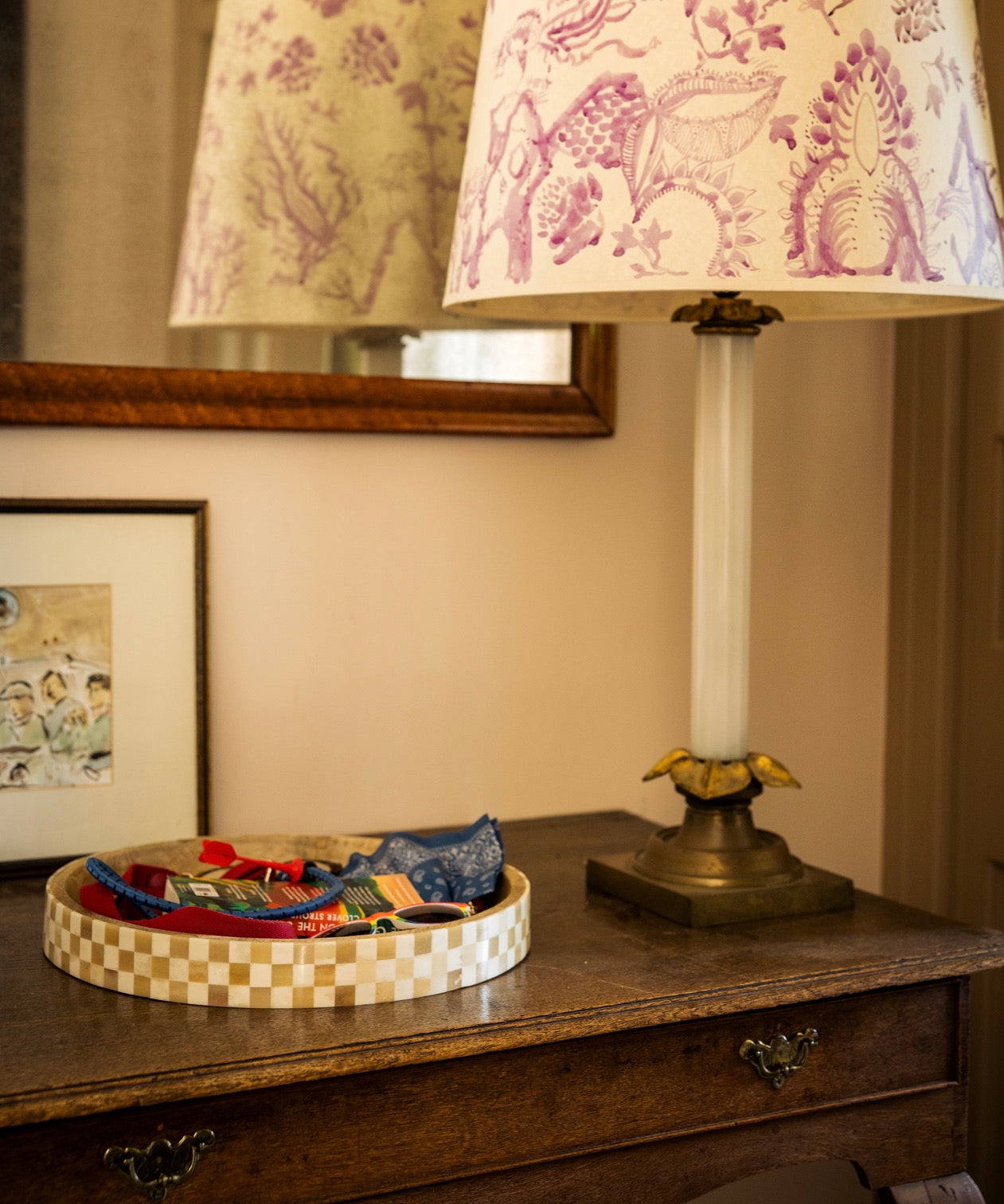 Decorative lamp with patterned shade on a console next to a natural wooden tray holding nicknacks, with a mirror on the wall in the background.