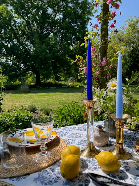 A blue and yellow tablescape with lemons, candlesticks, bud vases, flower arrangements, glasses, plates and placemats in the foreground. In the background is a pole with pink climbing roses, grass and a large tree.