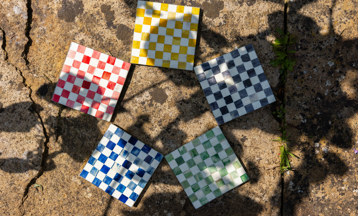 An overhead view of 5 coasters arranged in a circle, each one is a different colour checkerboard pattern on a brown stone background.