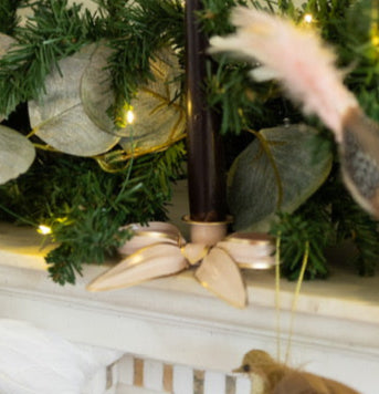 A close up of a pale pink metal bow candleholder with dark black wax taper candle on a mantlepiece in front of fairy lights, dried eucalyptus and pine branches.