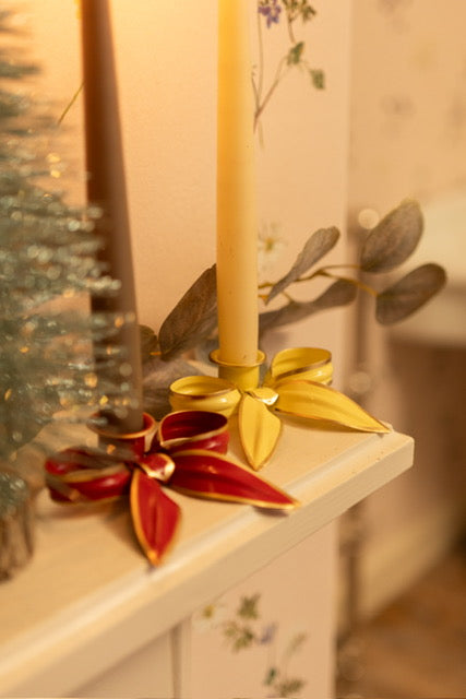 A mantlepiece with two metal bow candleholders one red with a grey candle the other yellow with an ivory candle.