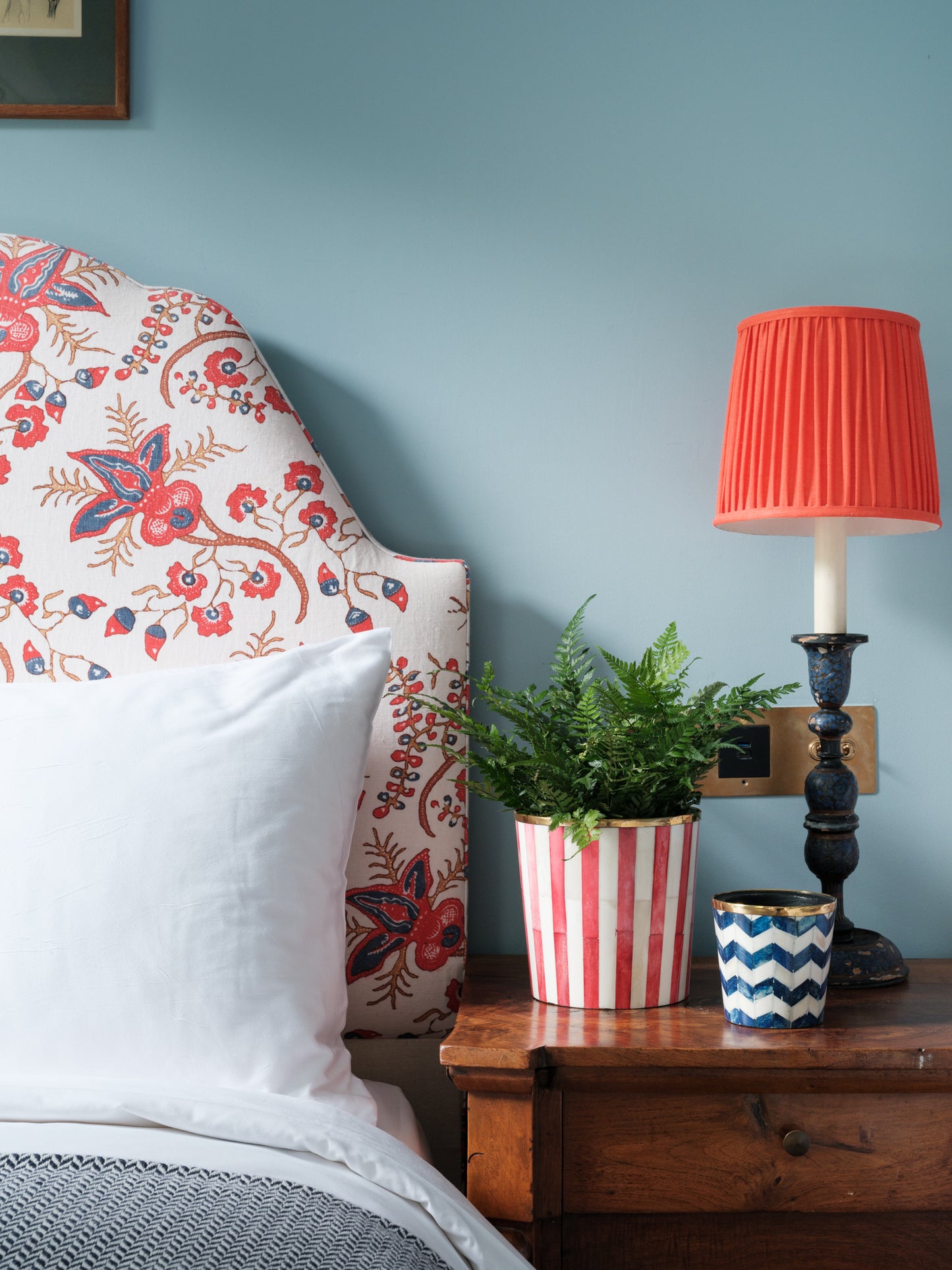 A wooden bedside table with fern stripe planter and small zigzag pot next to a red column lamp and floral headboard.