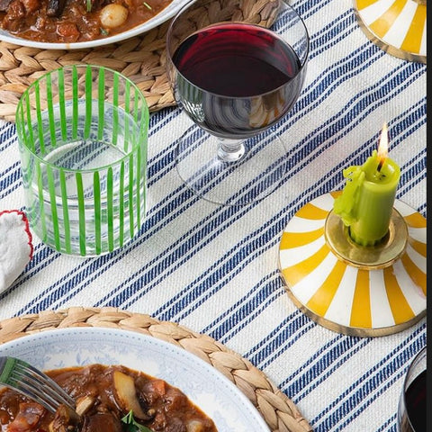 An overhead view of a table set for two with water tumbler, glass of red wine next to two round candleholders with burning green candles and two plates of food on straw placemats on a striped tablecloth. 