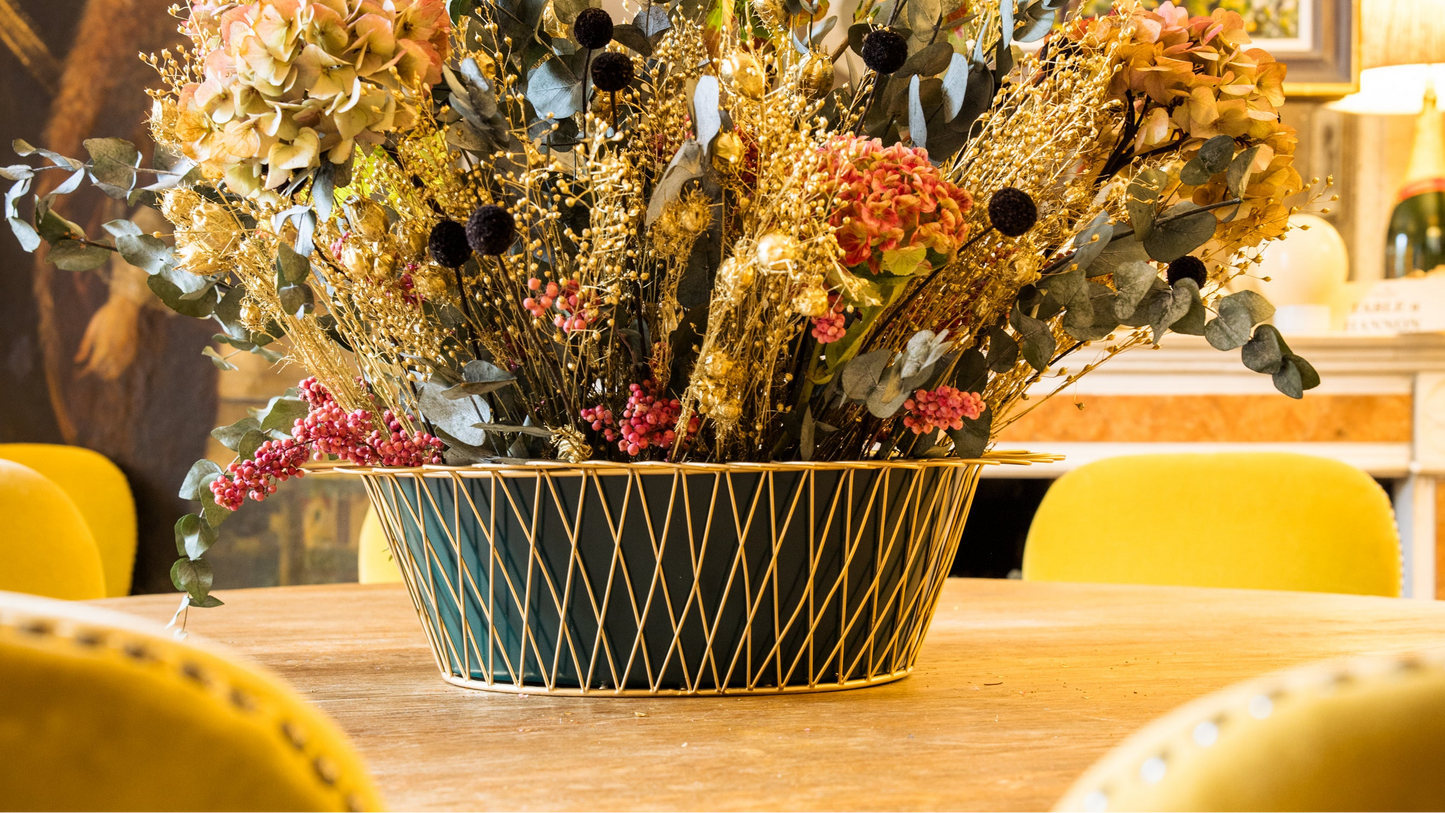 Decorative floral arrangement in a black and gold container on a table with yellow chairs.