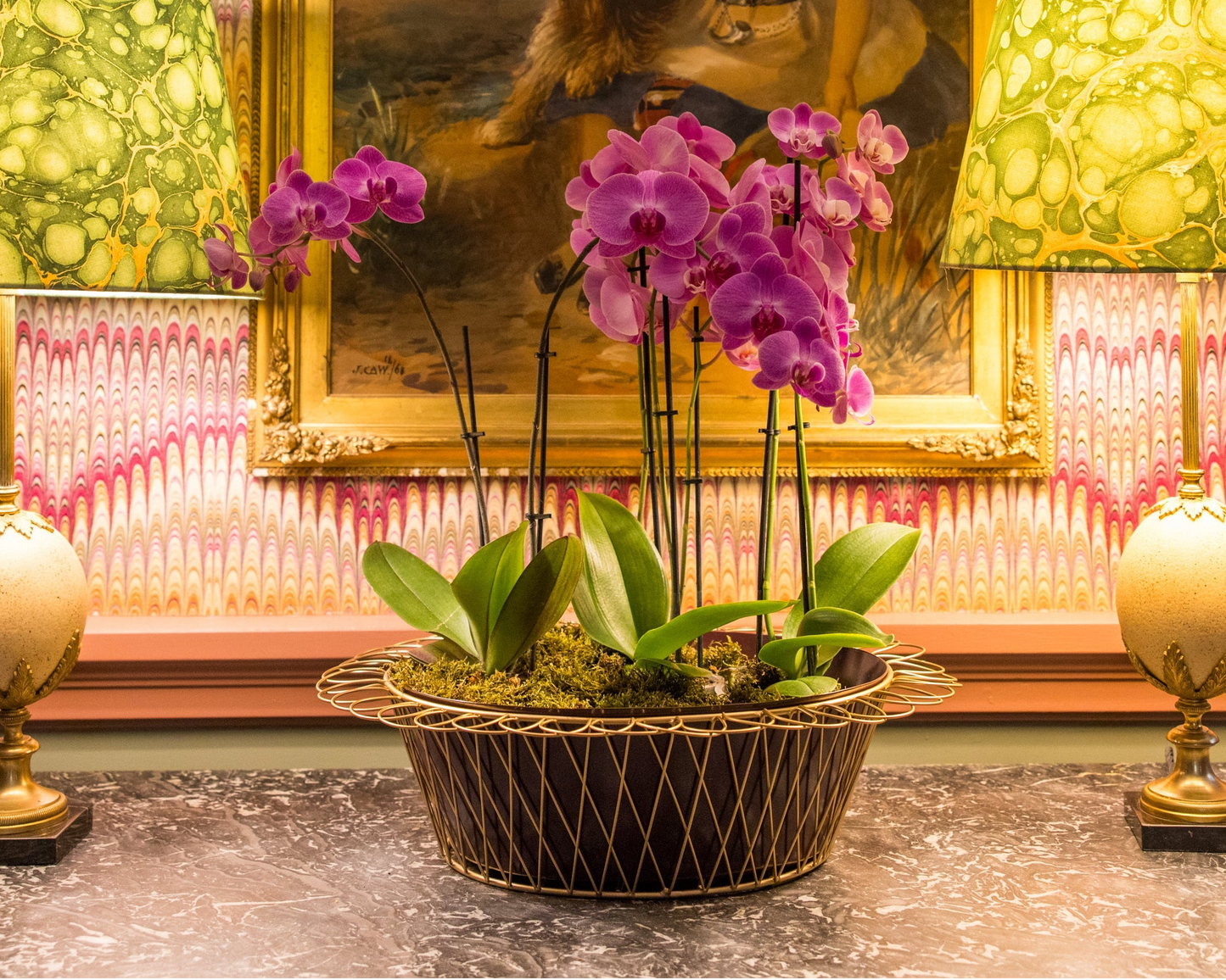 Purple orchids in a decorative pot on a table with a painting and lamps in the background.