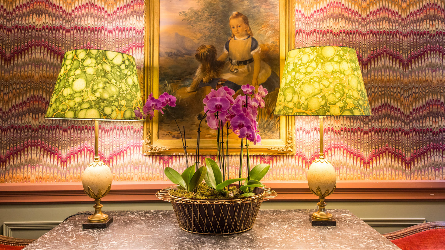 Decorative table setting with two lamps, a potted plant, and a framed portrait against a patterned wall.